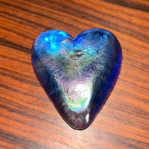 May include: A blue glass heart-shaped decorative object. The heart has a textured surface with iridescent highlights of purple, green, and blue. The object is resting on a wooden surface.