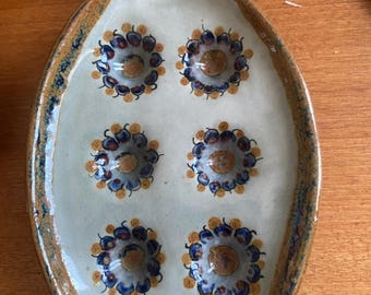 May include: Oval ceramic serving dish with a cream-colored interior and a brown rim. The dish features six raised floral designs in blue, brown, and gold. The dish is likely handmade and suitable for serving appetizers or small portions.