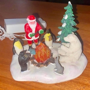 May include: A Christmas-themed figurine featuring Santa Claus, penguins, a polar bear, and a rabbit gathered around a campfire. A green Christmas tree and a white base complete the scene. The figurine is lit with a small light.