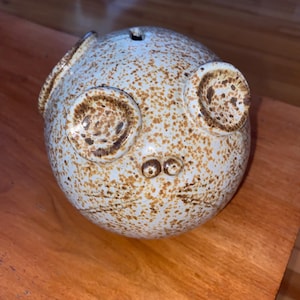 Könnte beinhalten: Ein Keramik-Sparschwein mit hellblauer Glasur und braunen Flecken. Das Sparschwein hat zwei große Ohren und zwei kleine Augen.