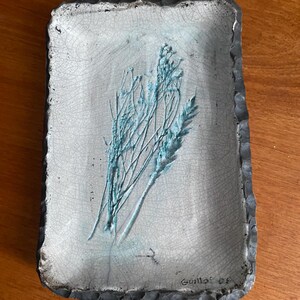 May include: Rectangular ceramic tray with a light blue and gray crackle glaze. The tray features a raised design of blue botanical elements. The edges are dark gray and textured. The artist's signature and date are visible in the bottom right corner.