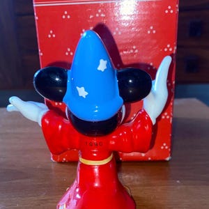 K&ouml;nnte beinhalten: Figur eines Zauberer-Charakters in roter Robe, blauem Hut mit wei&szlig;en Sternen und schwarzen Ohren. Die Figur hat wei&szlig;e Handschuhe und steht vor einer roten Schachtel mit dem Wort "Schmid". Das Jahr "1990" ist sichtbar.