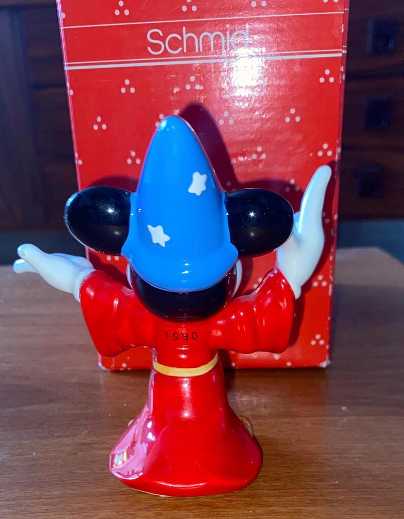 K&ouml;nnte beinhalten: Figur eines Zauberer-Charakters in roter Robe, blauem Hut mit wei&szlig;en Sternen und schwarzen Ohren. Die Figur hat wei&szlig;e Handschuhe und steht vor einer roten Schachtel mit dem Wort "Schmid". Das Jahr "1990" ist sichtbar.