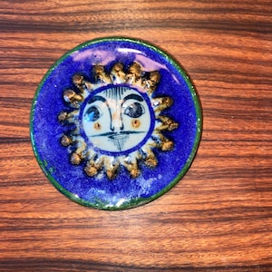 May include: A small, oval ceramic dish with a blue glaze and a green rim. The dish features a hand-painted sun with a face, surrounded by gold-coloured rays.