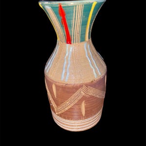 May include: A handcrafted ceramic vase with a flared top and a textured surface. The vase features a brown base with carved geometric patterns and a beige midsection with vertical lines. The top is teal with red and yellow accents.