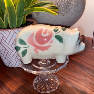 May include: White ceramic piggy bank with a pink rose and green leaves painted on the side. The piggy bank is sitting on a clear glass pedestal.