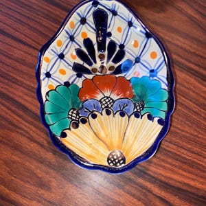 May include: A hand-painted ceramic dish with a scalloped edge. The dish is decorated with a floral pattern in shades of blue, green, yellow, orange, and red. The dish has a blue rim and a gold accent.