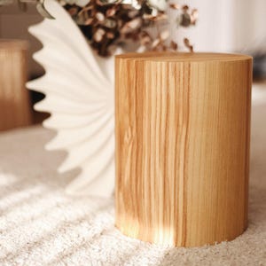 Op de afbeelding: Een cilindervormige houten bijzettafel met een lichtbruine afwerking. De tafel heeft een natuurlijk houtnerfpatroon en staat op een wit tapijt. Een witte vaas met een decoratief ontwerp en gedroogde bloemen staat op de achtergrond.