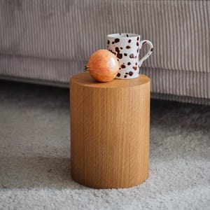 May include: A wooden side table with a pomegranate and a white mug with brown spots. A small brown dog sits on a grey corduroy sofa with fluffy cushions in the background. The table is cylindrical.