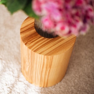 Op de afbeelding: Een cilindervormige houten kruk of bijzettafel met een lichtbruine afwerking en zichtbare natuurlijke houtnerven. Het bovenoppervlak is plat en rond en ondersteunt een donkere vaas gevuld met felroze hortensia's.