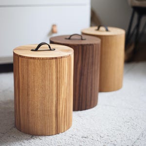 Könnte beinhalten: Drei Holzbehälter in verschiedenen Brauntönen. Jeder zylindrische Behälter hat einen Deckel mit einem schwarzen Griff. Die Behälter sind aus Holz gefertigt und haben ein natürliches Maserungsmuster.
