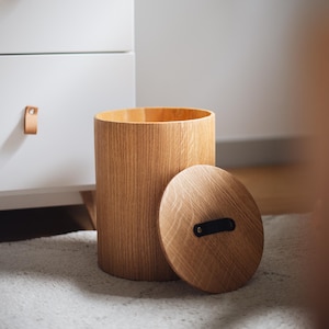 Op de afbeelding: Een cilindrische houten afvalbak met bijpassend deksel. De afvalbak is gemaakt van lichtgekleurd hout met een verticaal nerfpatroon. Het deksel heeft een zwarte leren handgreep. De afvalbak staat naast een witte ladekast met leren handgrepen.