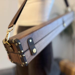 Puede incluir: Un estuche rectangular de madera con una correa de cuero marrón y herrajes dorados. El estuche tiene esquinas marrones oscuras con tachuelas doradas y un adorno de cuero. El estuche está diseñado para llevarse al hombro.