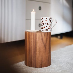 May include: A lit white candle and a speckled mug sit on a cylindrical wooden side table. The mug has a white background with brown spots. The table is made of dark wood. The scene is set in a room with a white cabinet.