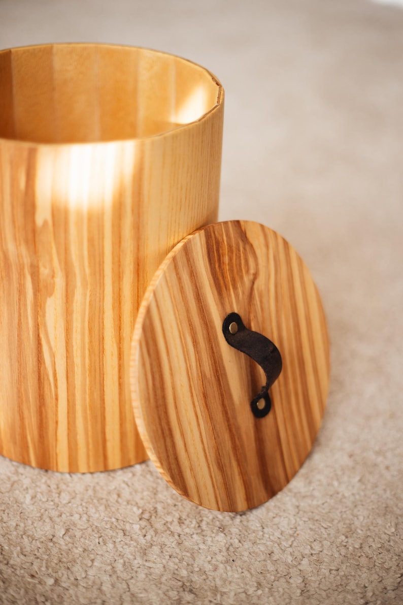 May include: A cylindrical wooden container with a matching lid. The container is made of light-colored wood with visible grain. The lid has a black leather handle. The container is likely for storage.