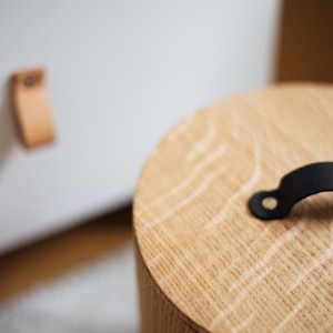 May include: A close-up of a wooden, cylindrical container with a black handle. The container has a light brown, natural wood grain finish. A white cabinet with a leather pull is slightly out of focus in the background.