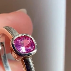 May include: A silver-toned ring featuring a cushion-cut, vibrant pink gemstone set in a smooth, rounded bezel setting. The band has a polished metallic finish, and the gemstone displays multiple facets that reflect light across its surface.