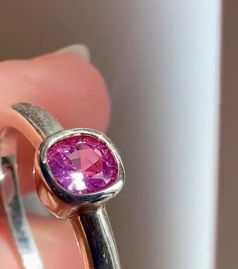 May include: A silver-toned ring featuring a cushion-cut, vibrant pink gemstone set in a smooth, rounded bezel setting. The band has a polished metallic finish, and the gemstone displays multiple facets that reflect light across its surface.