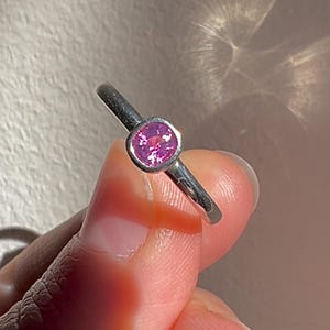 May include: A silver-toned ring featuring a single, round, faceted pink gemstone set in a smooth, bezel-style metal mounting. The band is plain and polished, showcasing a reflective metallic surface that catches the light.