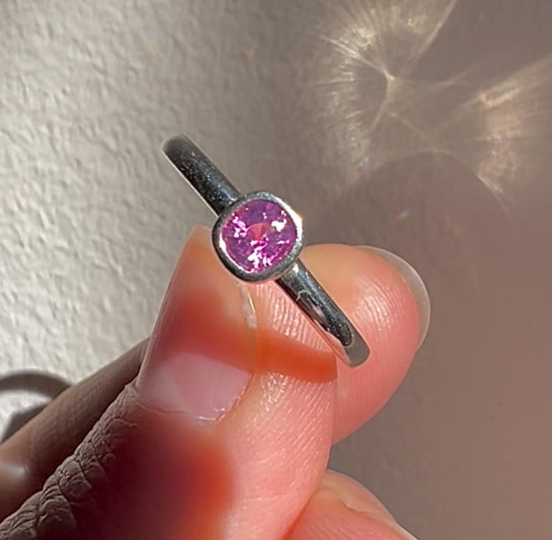 May include: A silver-toned ring featuring a single, round, faceted pink gemstone set in a smooth, bezel-style metal mounting. The band is plain and polished, showcasing a reflective metallic surface that catches the light.