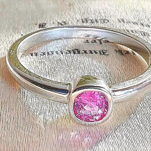 May include: A silver-toned metal ring featuring a single, round, faceted pink gemstone set in a smooth, bezel-style mounting. The band has a polished, reflective finish and a simple, rounded profile that encircles the central pink stone.