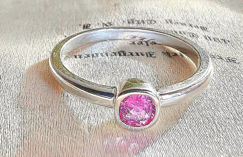 May include: A silver-toned metal ring featuring a single, round, faceted pink gemstone set in a smooth, bezel-style mounting. The band has a polished, reflective finish and a simple, rounded profile that encircles the central pink stone.