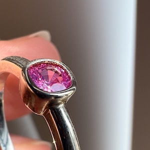 May include: A ring featuring a faceted, cushion-cut pink gemstone set in a smooth, polished silver-toned metal bezel. The band is rounded and metallic, showcasing a reflective surface that catches the light around the central pink stone.