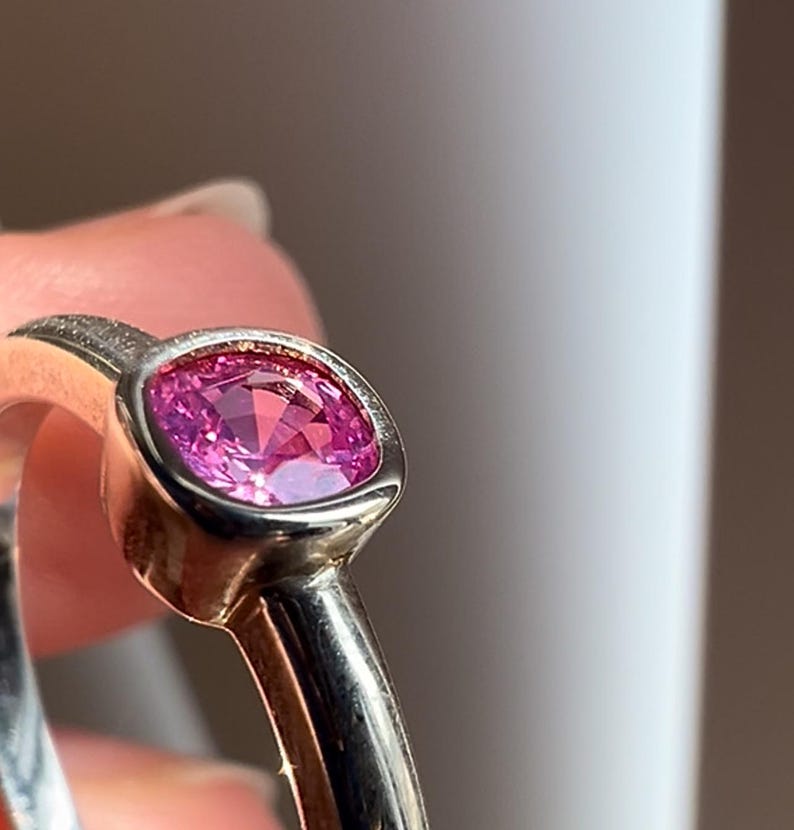 May include: A ring featuring a faceted, cushion-cut pink gemstone set in a smooth, polished silver-toned metal bezel. The band is rounded and metallic, showcasing a reflective surface that catches the light around the central pink stone.
