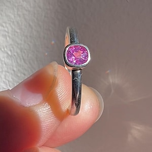 May include: A silver ring featuring a single, faceted, cushion-cut pink gemstone set in a smooth, polished bezel setting. The band is plain and metallic, showcasing a reflective surface that catches the light, highlighting the vibrant pink stone.