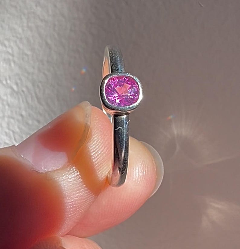 May include: A silver ring featuring a single, faceted, cushion-cut pink gemstone set in a smooth, polished bezel setting. The band is plain and metallic, showcasing a reflective surface that catches the light, highlighting the vibrant pink stone.