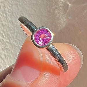 May include: A silver-toned ring featuring a single, cushion-cut, vibrant pink gemstone set in a smooth, rounded bezel setting. The band is polished and metallic, showcasing a clean, simple design that highlights the bright pink center stone.