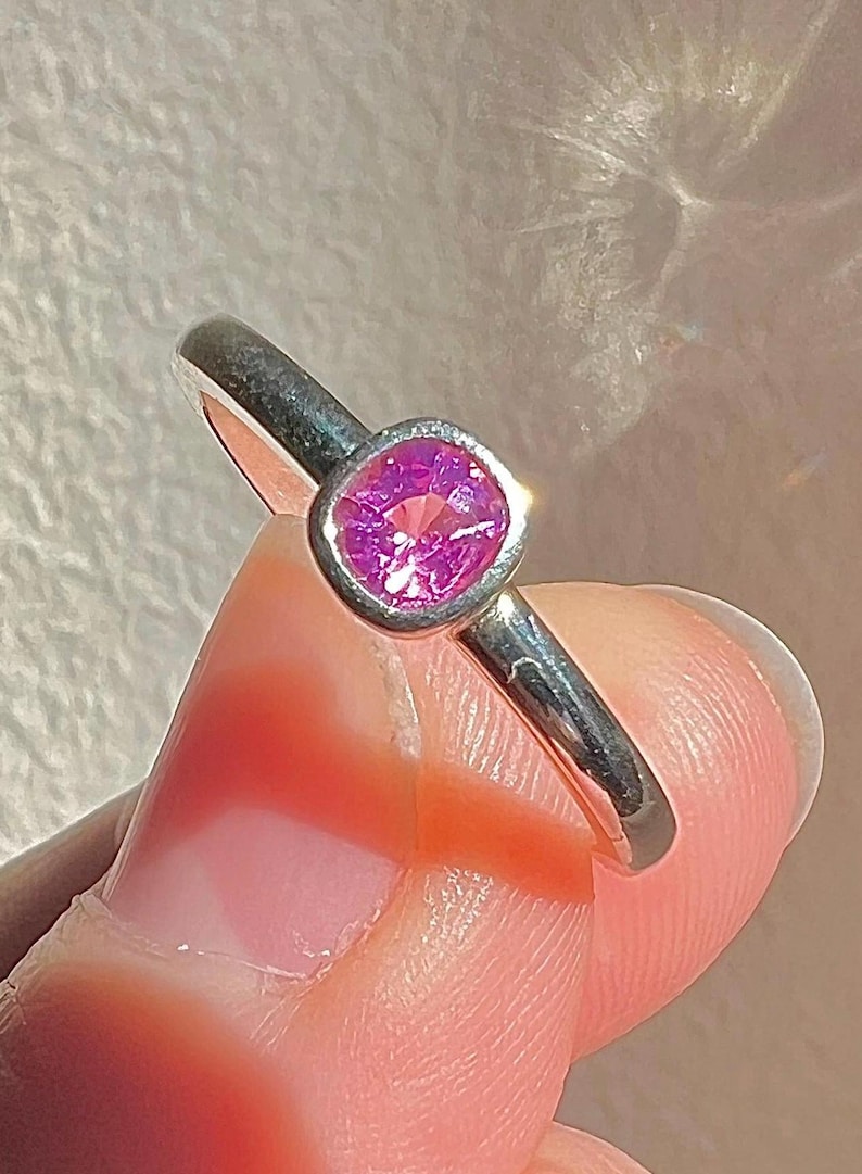 May include: A silver-toned ring featuring a single, cushion-cut, vibrant pink gemstone set in a smooth, rounded bezel setting. The band is polished and metallic, showcasing a clean, simple design that highlights the bright pink center stone.