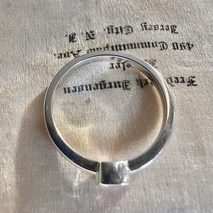 May include: A polished silver ring with a smooth, rounded band and a prominent, rectangular setting at the base. The metal surface reflects light, showing a clean, metallic finish. The ring is positioned over text that reads Frederick Jurgensen 490 Communipaw Ave. Jersey City, N.J.