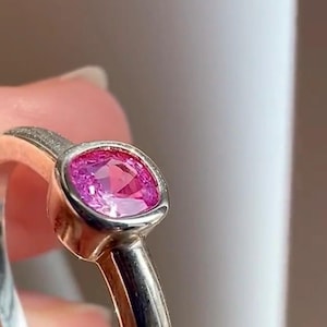May include: A ring featuring a cushion-cut vibrant pink gemstone set in a smooth, polished silver-toned metal band. The setting surrounds the faceted stone, highlighting its deep pink color and reflective surface within the metallic frame.