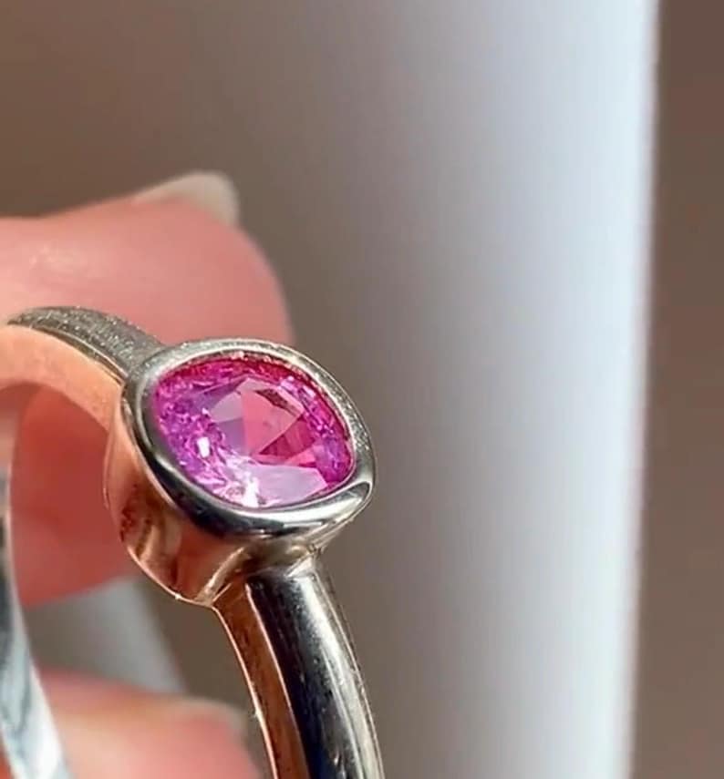 May include: A ring featuring a cushion-cut vibrant pink gemstone set in a smooth, polished silver-toned metal band. The setting surrounds the faceted stone, highlighting its deep pink color and reflective surface within the metallic frame.