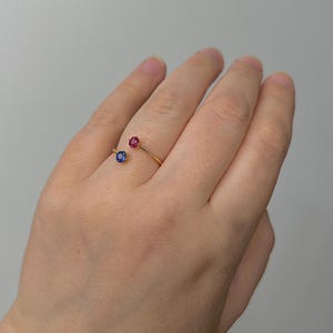 Könnte beinhalten: Ein goldener Ring mit einem roten und blauen Edelstein. Der Ring ist offen und umschließt den Finger.