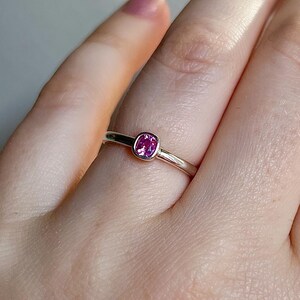 May include: A silver-toned ring featuring a single, small, cushion-cut pink gemstone set in a smooth bezel setting. The band is polished and metallic, with a simple, rounded profile that encircles the finger, highlighting the vibrant pink stone.