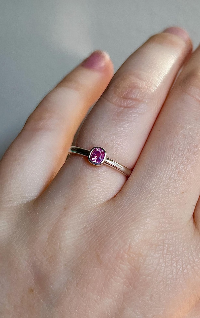 May include: A silver-toned ring featuring a single, small, cushion-cut pink gemstone set in a smooth bezel setting. The band is polished and metallic, with a simple, rounded profile that encircles the finger, highlighting the vibrant pink stone.