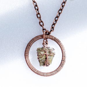 Op de afbeelding: Handgemaakte koperen ketting met een groene kristallen hanger. De hanger hangt in een koperdraadcirkel, met een koperen ketting. Het ontwerp bevat een levensboommotief, dat een organische toets aan het sieraad toevoegt.