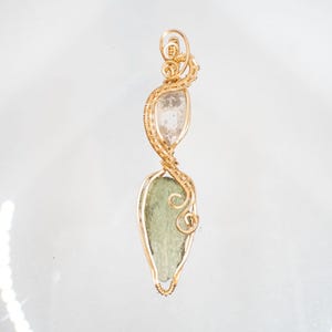 May include: A handcrafted pendant featuring a clear, oval-shaped crystal above a green, leaf-shaped stone, both encased in gold-colored wire. The wire is intricately woven, creating a decorative, swirling design. The pendant is suspended from a loop at the top.