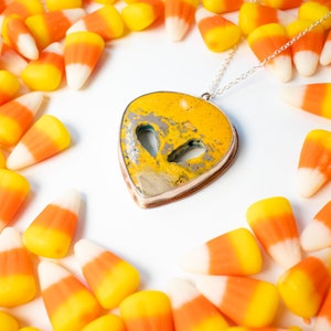 May include: A silver chain necklace with a yellow and brown heart-shaped pendant. The pendant has a unique marbled pattern and is surrounded by candy corn.