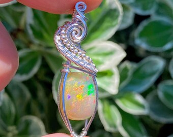 May include: A silver wire wrapped pendant featuring a colorful opal gemstone. The pendant has a delicate, swirling design and is perfect for adding a touch of sparkle to any outfit.