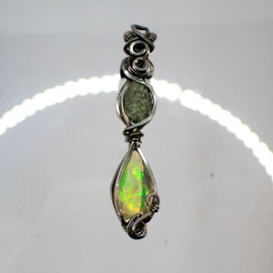 Puede incluir: Colgante artesanal con diseño de alambre plateado. Presenta dos piedras preciosas: una piedra ovalada verde y un ópalo en forma de pera con vibrantes tonos verdes y amarillos. El trabajo de alambre añade un toque decorativo a esta pieza de joyería única.