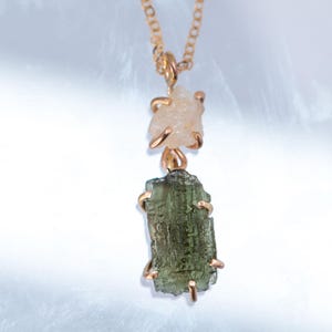 May include: A gold-toned necklace featuring two raw, uncut gemstones suspended in a vertical arrangement. The top stone is a pale, translucent peach colour, while the bottom stone is a larger, textured, deep olive-green rectangular crystal.