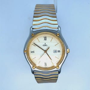 Op de afbeelding: Een tweekleurig Ebel-polshorloge met een zilveren en gouden golvende schakelarmband, een ronde zilveren kast met gouden schroeven en een crèmekleurige wijzerplaat met Romeinse cijfers, gouden wijzers en een datumvenster met het getal 19.
