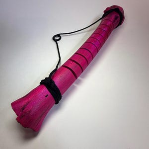 May include: A cylindrical wooden object painted in a vibrant magenta hue with a textured, natural wood grain finish. The surface features a series of evenly spaced horizontal grooves. A black cord is wrapped around the base and top ends.