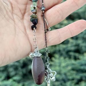 May include: A handmade pendant featuring a dark brown oval stone, a chain of colorful gemstones, and a silver fairy charm. The pendant also includes a chain of star-shaped charms and a silver clasp.