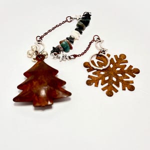 May include: Two Christmas-themed charms. One is a rusty brown tree, the other a rusty brown snowflake. Both are connected by a chain with a mix of beads and stars. The charms are on a white background.