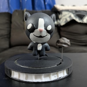 May include: A black and white cartoon cat figurine with a pink nose and a small rock on a metal stand. The cat is standing on a black circular base with a white honeycomb design.