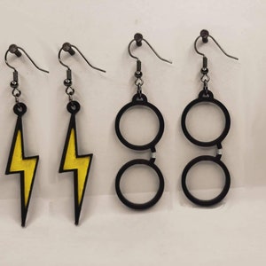 May include: Four pairs of earrings. Two pairs feature a yellow lightning bolt design with black outlines. The other two pairs are black-rimmed glasses. All earrings have black metal hooks and small beads.